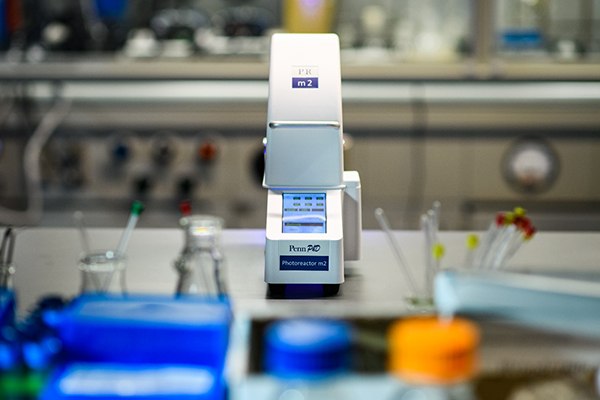 A close-up view of the Penn PhD Photoreactor M2, a laboratory instrument with a sleek white design and a digital display, positioned on a lab countertop surrounded by various laboratory glassware and equipment, including colorful reagent bottles and pipettes in the background. A close-up view of the Penn PhD Photoreactor M2, a laboratory instrument with a sleek white design and a digital display, positioned on a lab countertop surrounded by various laboratory glassware and equipment, including colorful reagent bottles and pipettes in the background.
