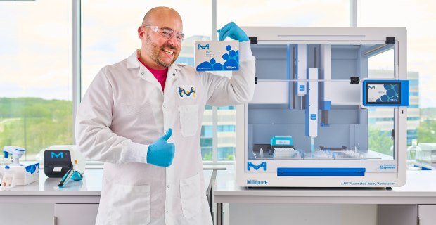 Scientist standing in front of AAW™ automated assay workstation holding up SMC® assay giving a thumbs up. Scientist standing in front of AAW™ automated assay workstation holding up SMC® assay giving a thumbs up.